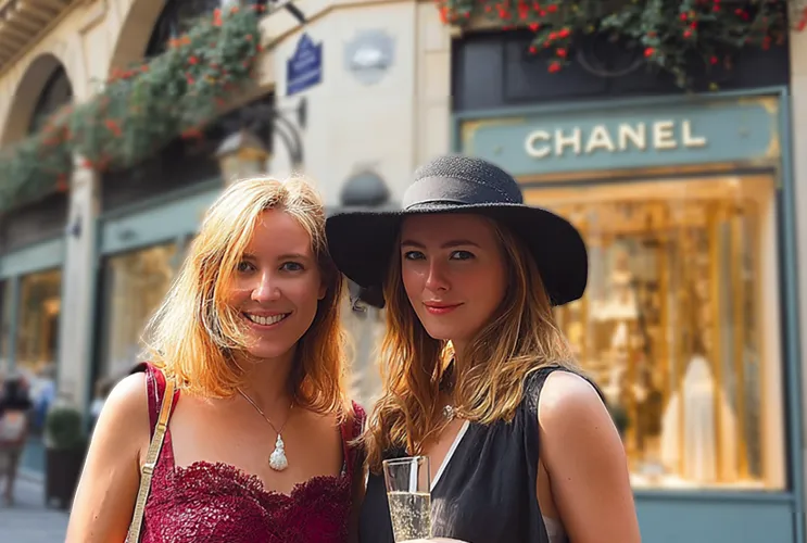 Two women in Paris, one holding a champagne glass, in front of a Chanel boutique — Paris Fashion Tour.