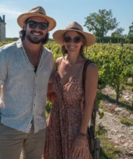 Smiling couple wearing summer hats in Bordeaux vineyards with a château in the background, Saint-Émilion wine region